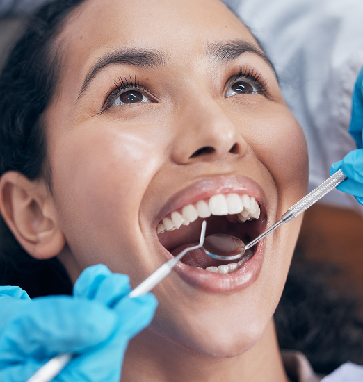 A woman smiles during a dental checkup, with gloved hands holding a mirror and probe in her mouth. The mood is positive and relaxed.