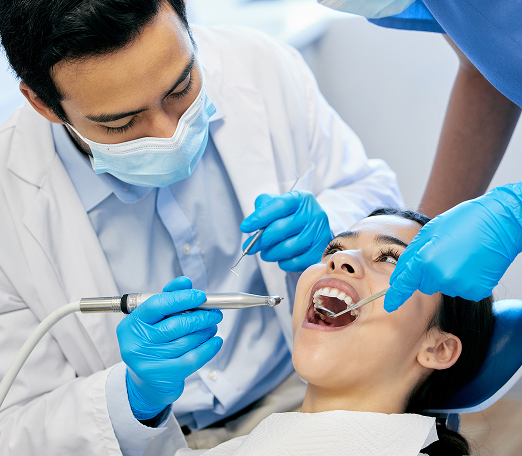 Dentist in a white coat examines a patient's teeth using dental tools. The patient is reclined, wearing a bib. Both wear blue surgical masks and gloves.