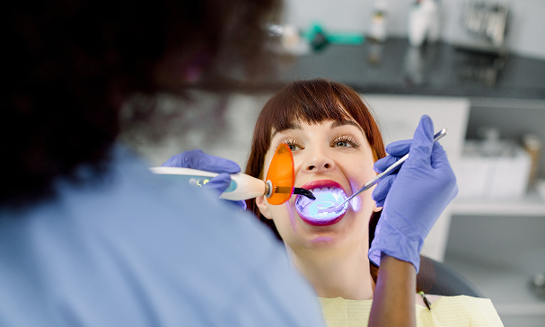 A dentist wearing gloves uses a curing light on a patient's teeth in a dental clinic. The patient is reclining comfortably, conveying a calm atmosphere.