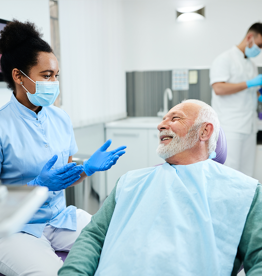 Dentist in a blue uniform and mask talks to a smiling senior man in a dental chair. Another dentist works in the background. Clinic setting, cheerful mood.