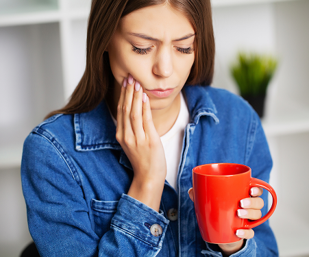 A woman in a denim jacket holds a red mug while touching her cheek, appearing to be in pain, possibly from a toothache. The background is softly blurred.