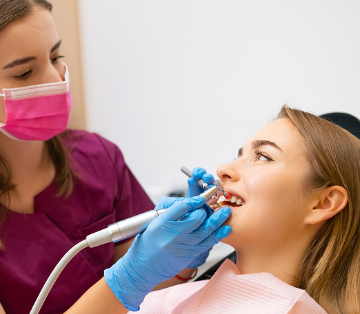 Dentist in pink mask and purple scrubs works on a patient's teeth using dental tools. The patient looks relaxed and the environment is clinical.