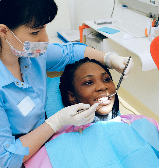 A dentist in a blue coat and patterned mask examines a smiling patient. The patient, wearing a blue bib, appears relaxed in a bright dental office.