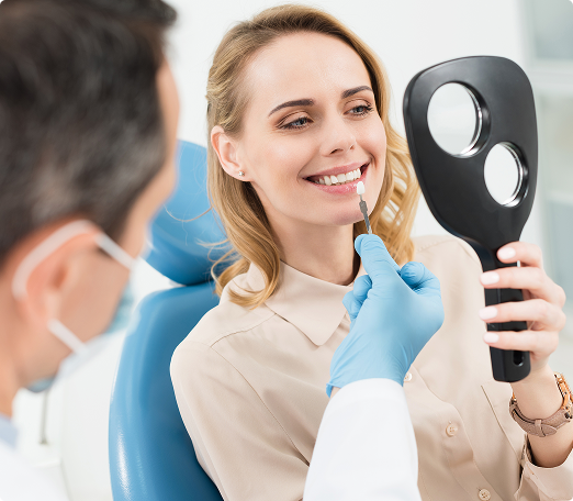 A woman in a dental chair smiles while holding a mirror. A dentist in gloves holds a tooth shade guide to her teeth. The atmosphere is professional and reassuring.