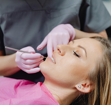 A dentist in gray scrubs and pink gloves examines a patient's teeth with a tool. The patient, wearing a pink bib, looks relaxed and calm.