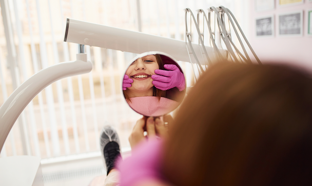 A woman at the dentist holds a round mirror, smiling at her reflection. She wears pink gloves, suggesting a playful, positive mood.
