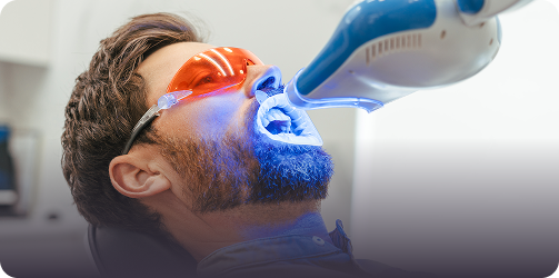 A man in orange protective glasses receives teeth whitening treatment with a blue light in a dental clinic, conveying a sense of focus and care.