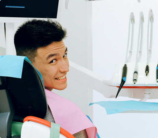 A person sits in a dental chair, smiling back at the camera. Dental tools are visible in the background, conveying a clinical but friendly atmosphere.