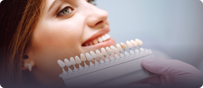 A woman smiles warmly as she holds a tooth shade guide near her mouth, wearing a pink glove. The scene conveys a sense of care and professionalism.