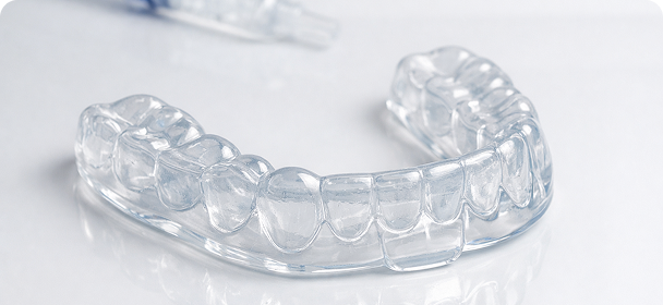 Clear dental aligner on a white surface, showcasing its transparent design and teeth structure. A dental tool is blurred in the background.