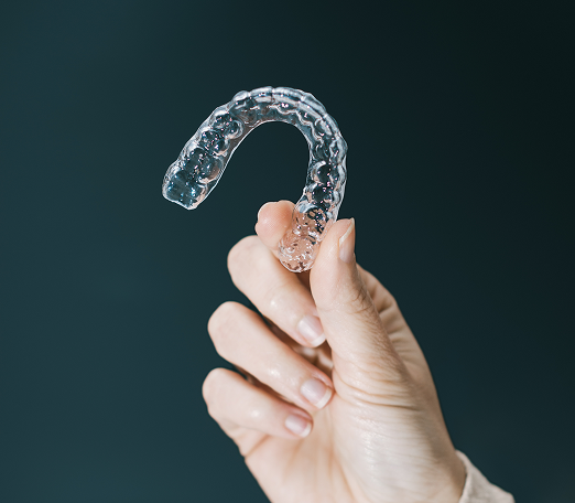 A hand holds a clear dental aligner against a dark background. The aligner is transparent and U-shaped, symbolizing dental care and orthodontics.